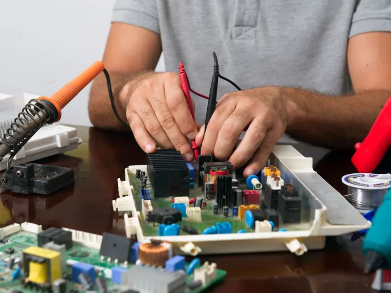 Man working on an electronic circuit board with tools on a table, showing close-up of hands and components.
