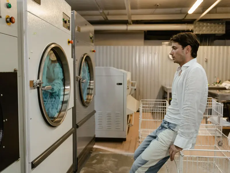 A man sits in a laundromat next to industrial washing machines, waiting for his laundry.