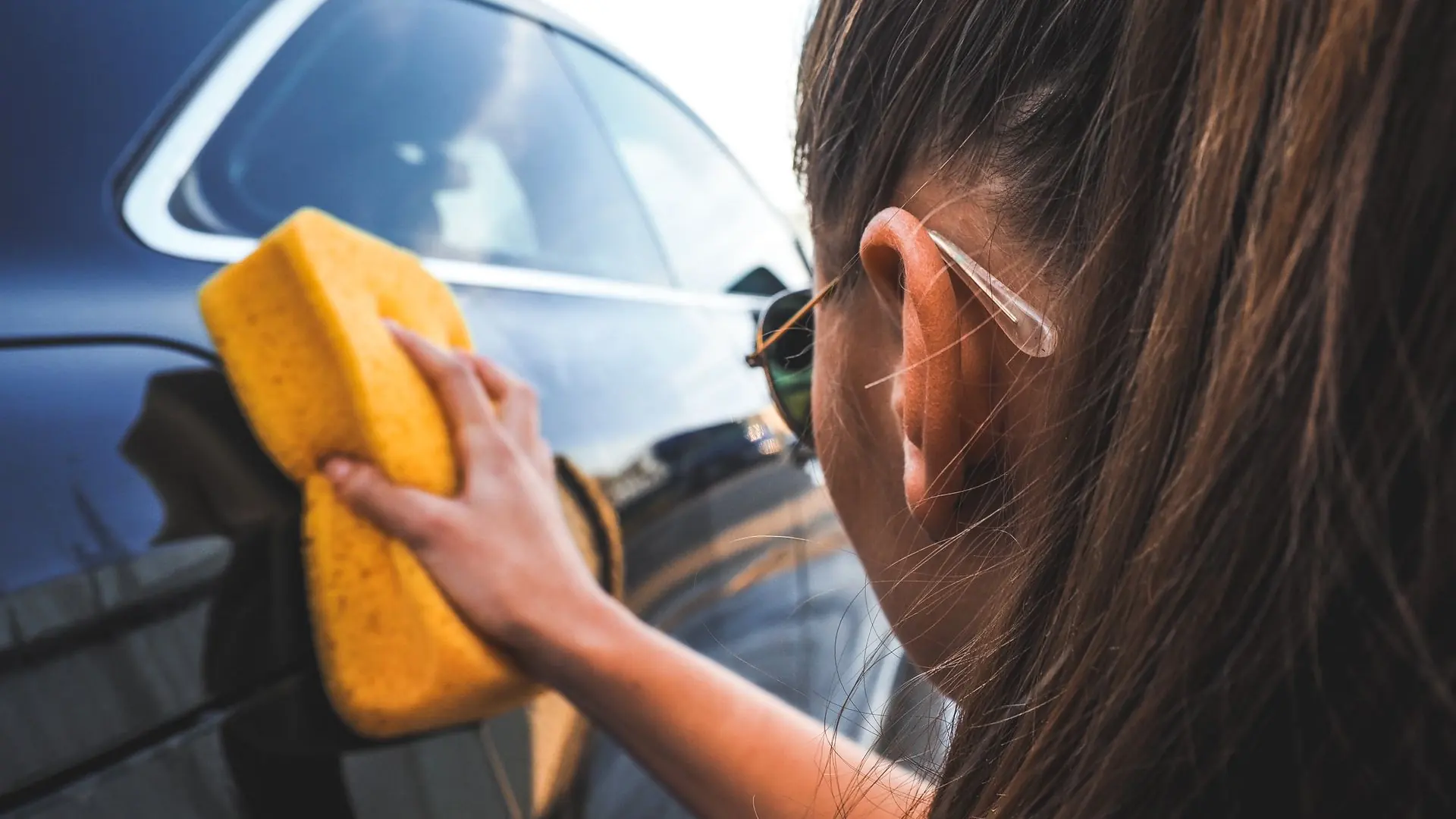 Close-up of a woman handwashing a black car using a yellow sponge, highlighting cleaning process.