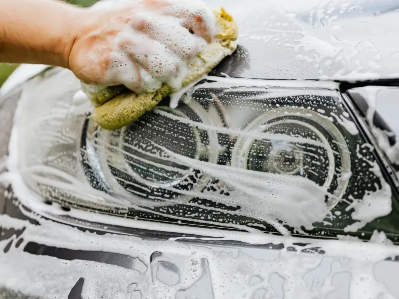 Hand scrubbing a car headlight with sponge and soap suds, detailing process.