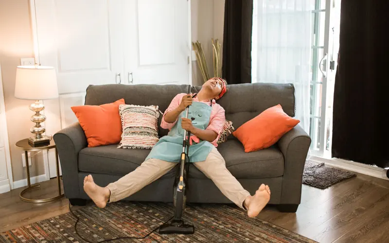 Man exhausted after vacuuming, resting on a couch with a vacuum cleaner at home.
