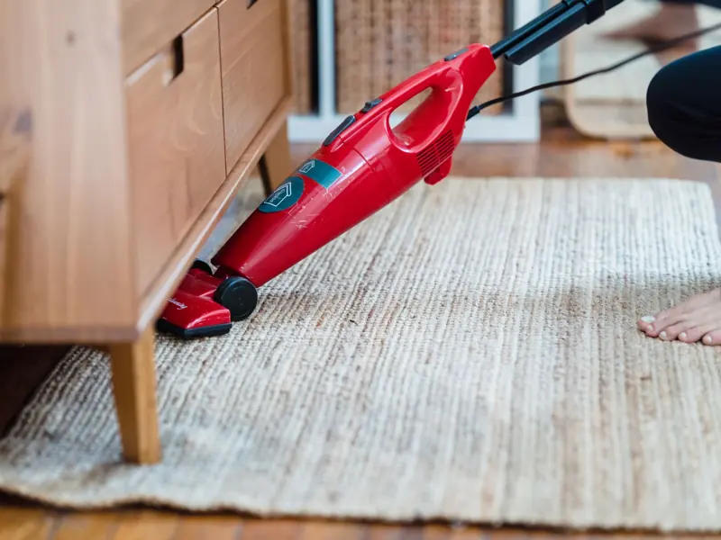 Side view of a person using a red vacuum under furniture on a rug for effective cleaning.