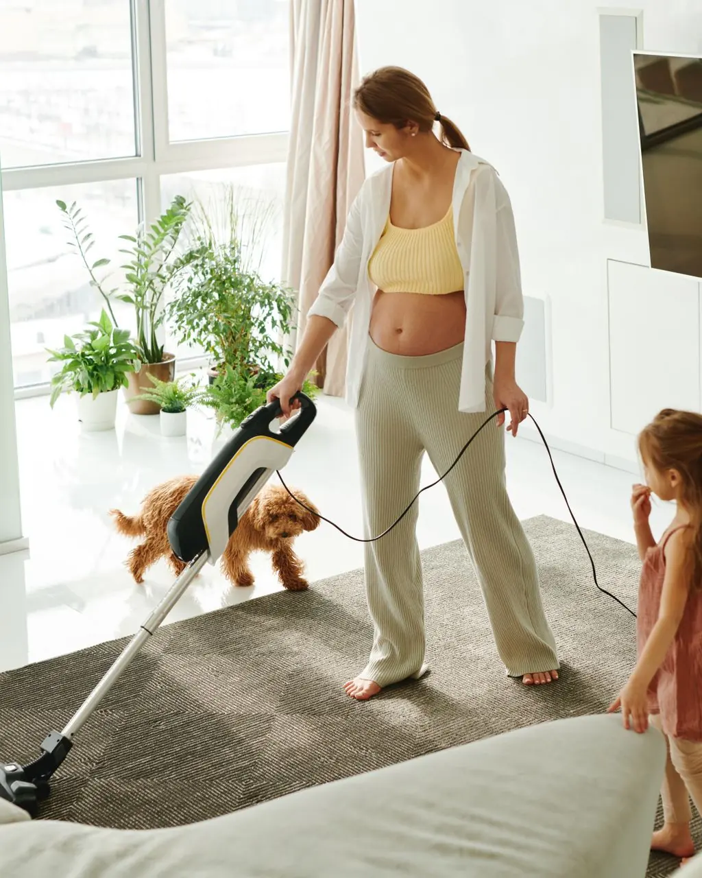 Pregnant mother cleaning carpet with vacuum as daughter and dog play indoors.