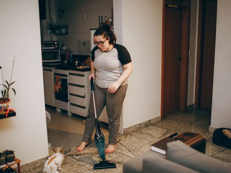 A woman cleaning her living room with a vacuum cleaner while her pet rabbit watches.