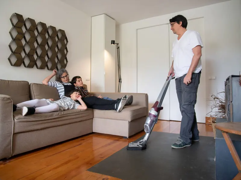A family relaxes on a sofa while a man vacuums the living room floor, capturing a moment of daily life.