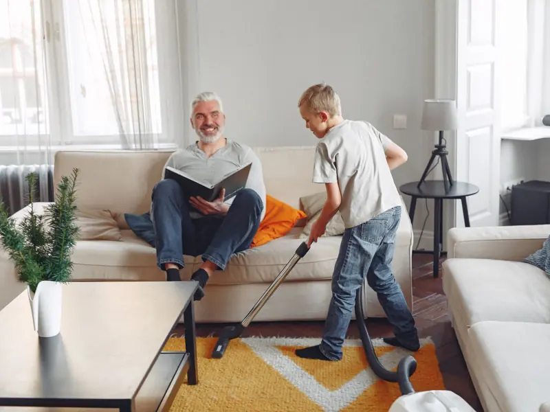 A father relaxing while his son vacuums in a cozy living room setting, showcasing family bonding.