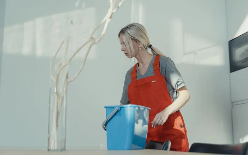 A woman in a red uniform cleans indoors with a blue bucket, ensuring a spotless environment.