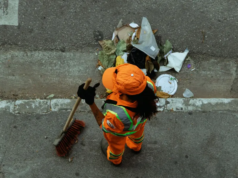 Top view of a street cleaner wearing orange uniform sweeping debris on a city sidewalk.