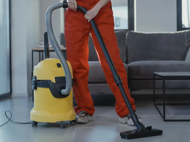 A professional cleaner in red coveralls vacuums a contemporary living room with a yellow vacuum cleaner.