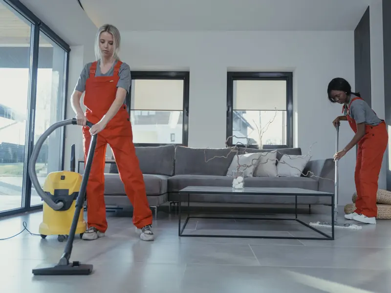 Two women in red overalls vacuuming and sweeping a modern living room.
