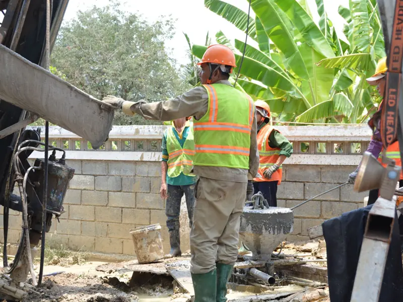 Construction workers in safety gear managing a cement mixer, surrounded by greenery.