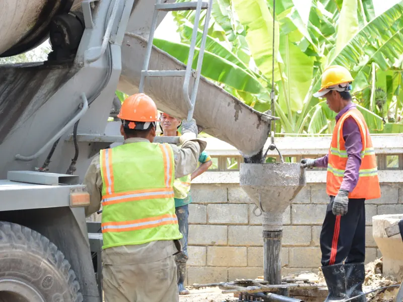 Construction workers managing cement pour from a mixer on site, wearing safety gear outdoors.