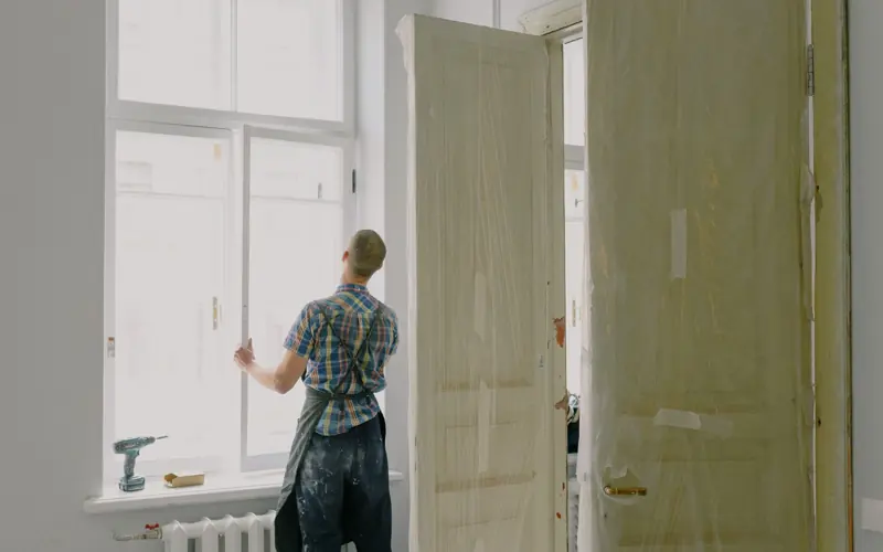 Back view of unrecognizable workman in apron mounting window near drilling machine and doors in flat