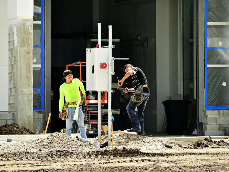 Two construction workers install an electrical panel outside a building site, wearing safety gear.