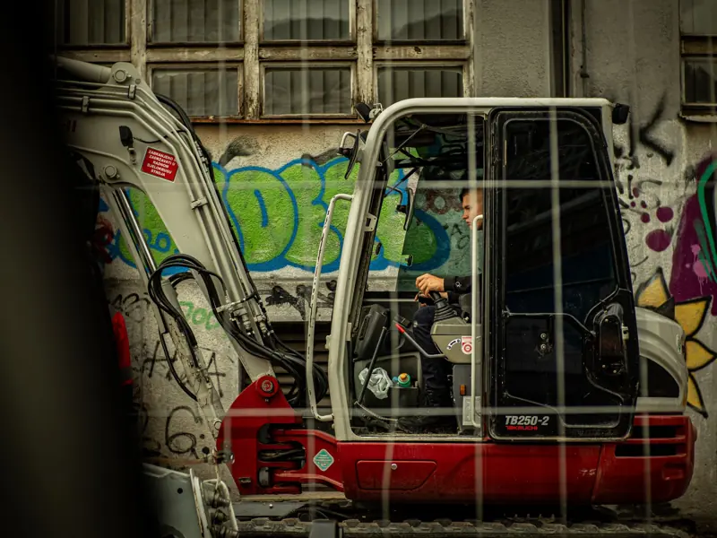 Man operating construction digger in urban setting with graffiti background.