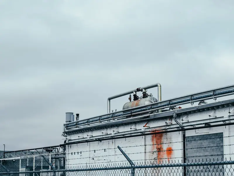 Shabby wall of factory with metal pipes and special equipment located near fence in industrial district of town against cloudy sky