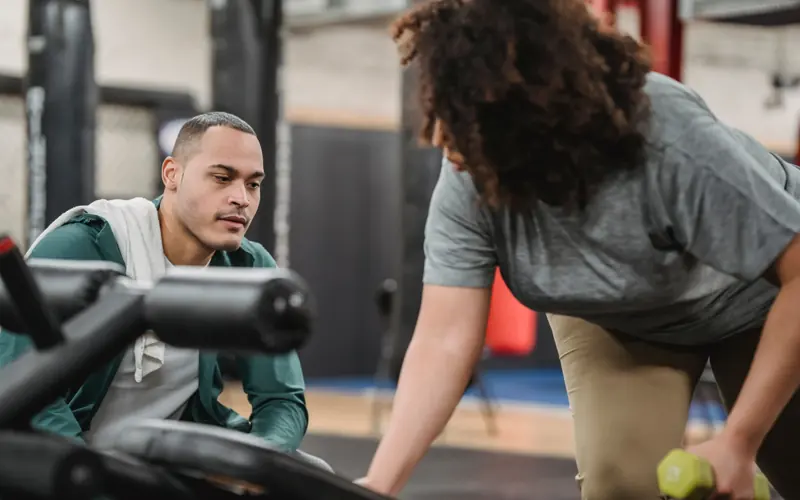 Experienced instructor watching anonymous African American female lifting dumbbell on blurred background of gym