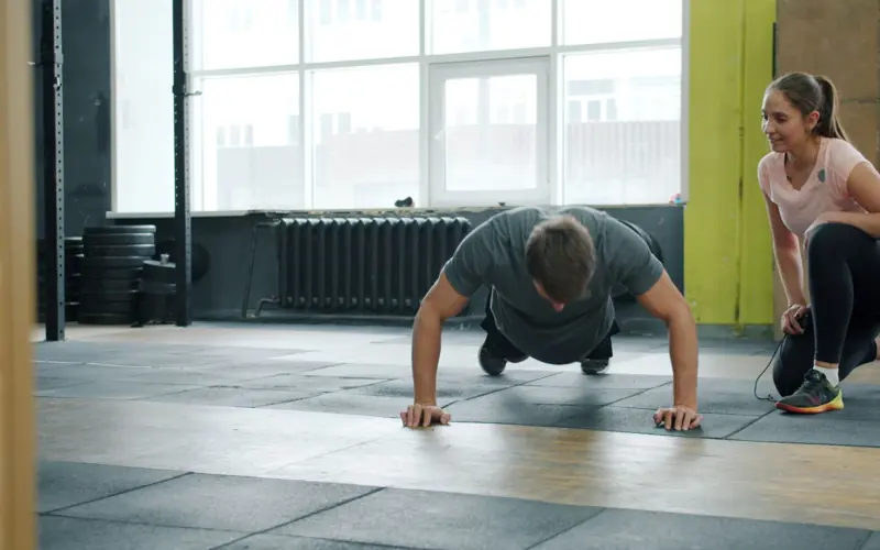 Man doing push-ups under supervision of a personal trainer in a gym setting.