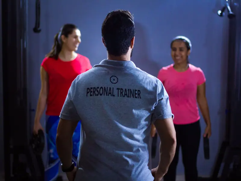 Back view of a personal trainer guiding two women during a gym session indoors.