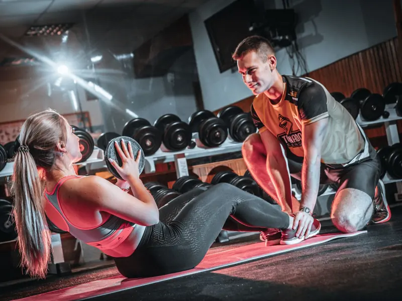 A personal trainer assists a woman doing sit-ups with a medicine ball in a gym setting.