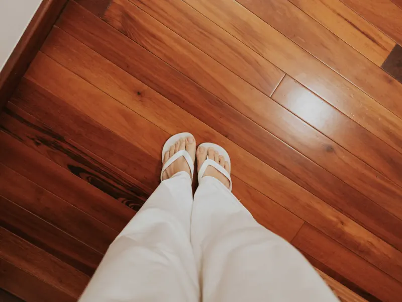 View of person wearing flip-flops and white pants standing on a wooden floor.