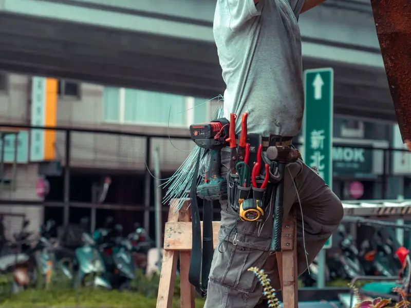 Handyman standing on a ladder with tool belt, repairing outdoors with visible tools.