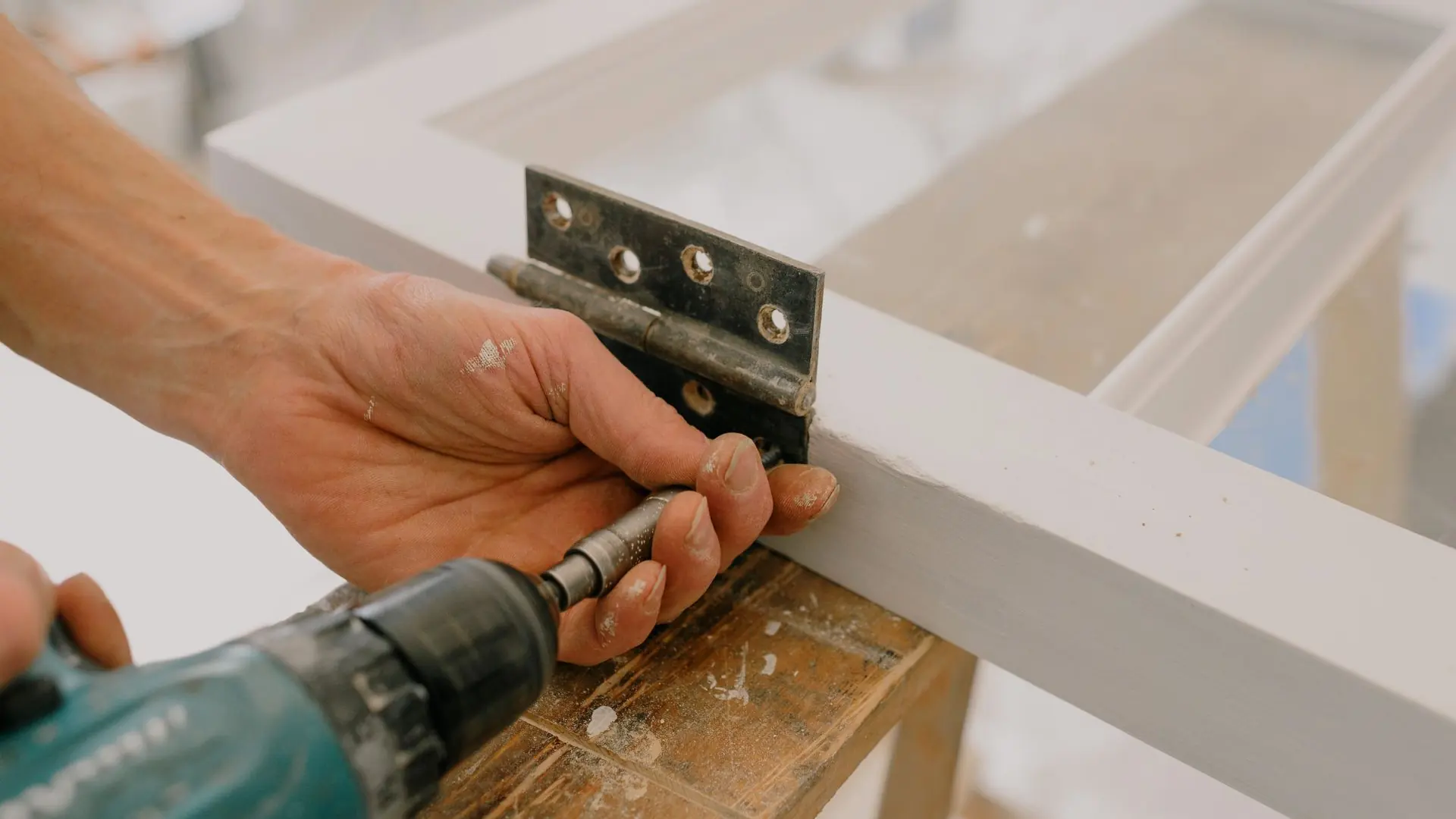 Crop unrecognizable worker drilling metal hinge on white window frame during house renovation