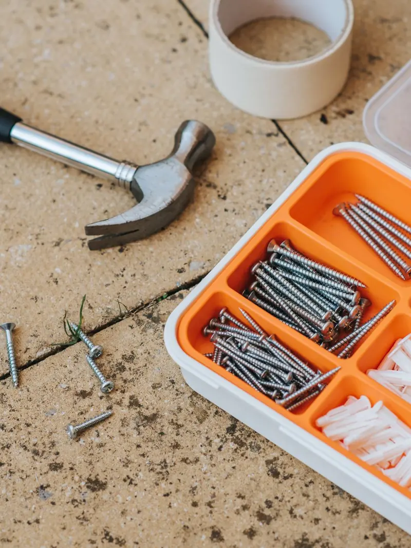 Top view of assorted repair instruments and screws in box near hammer and scotch tape on dirty tile in soft daylight