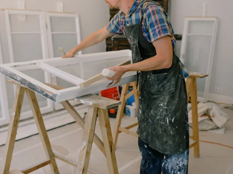 Unrecognizable male master wearing dirty apron standing near window with white frames on wooden workbench while working in professional studio
