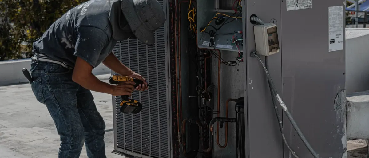 A worker in a bucket hat repairs an outdoor air conditioning unit on a rooftop.
