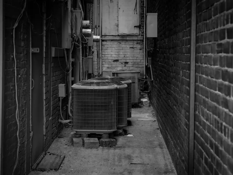 Black and white alley featuring industrial air conditioning units against a brick wall.