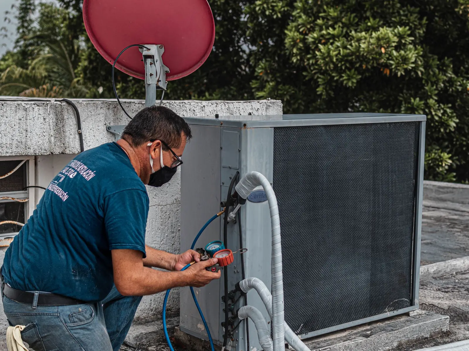 Technician repairing an air conditioner unit outdoors, wearing a facemask and using a manifold gauge.