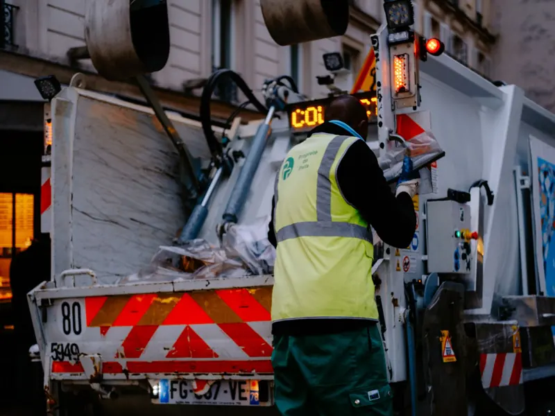 A garbage collector works on a city street, managing waste collection at dusk.