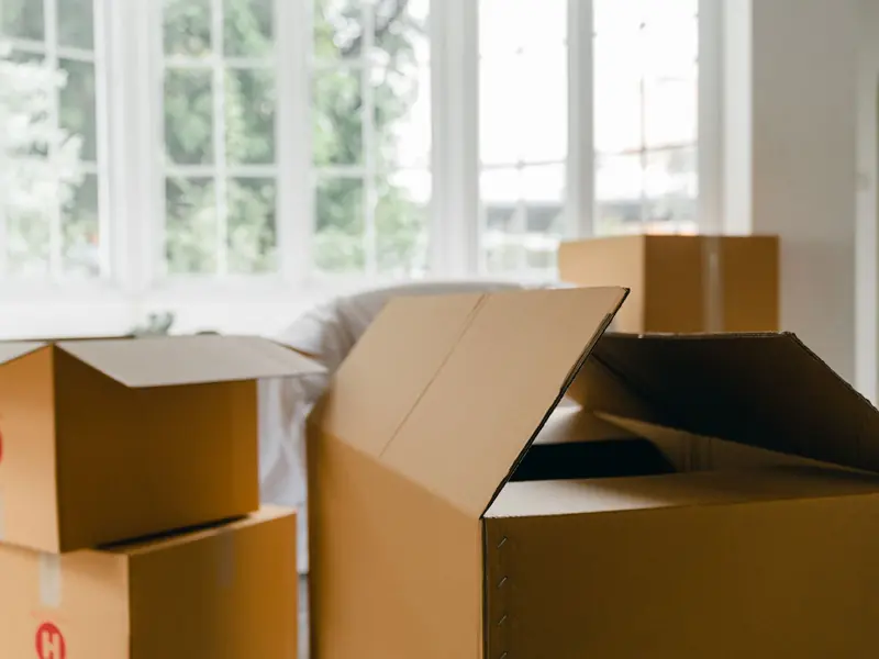 Empty unpacked carton boxes for moving personal items placed in middle of modern room near covered with sheet arm chair against panoramic window