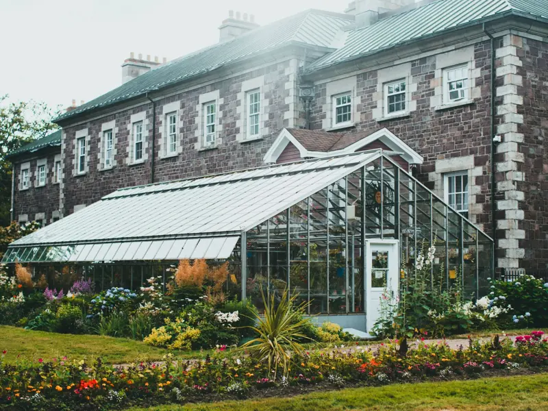 A picturesque greenhouse surrounded by vibrant gardens against a historic stone building.