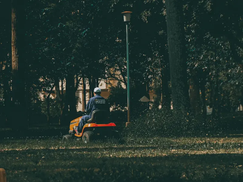 A man in coveralls operates a lawn mower in a verdant park setting, surrounded by lush trees and grass.