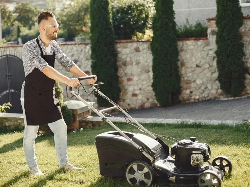 A man using a lawn mower to trim grass in a sunny outdoor garden.