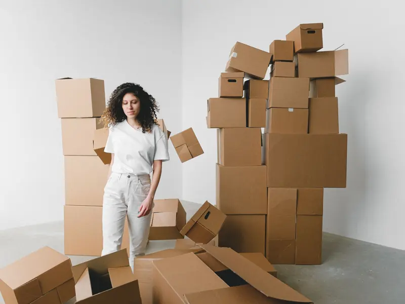 Young female with curly hair between piles and flying cardboard containers after moving into house on light background