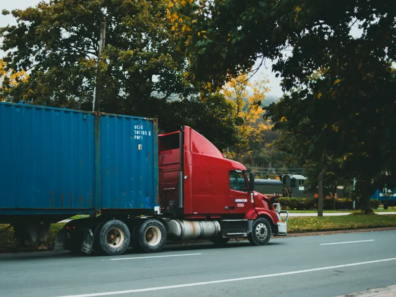 A red truck transports a blue cargo container on a scenic, tree-lined road.
