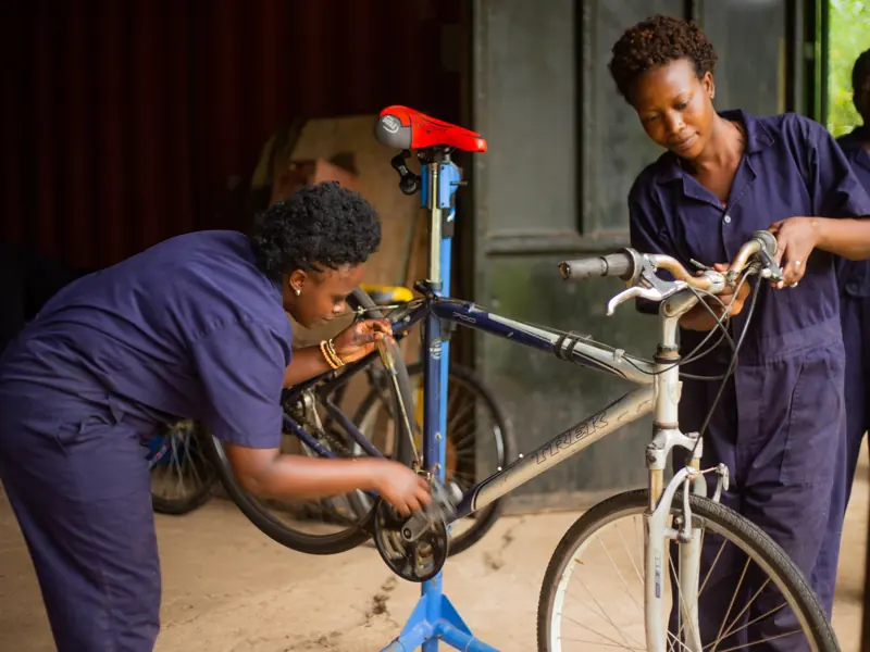 Two female mechanics in blue overalls working on a bicycle inside a workshop.