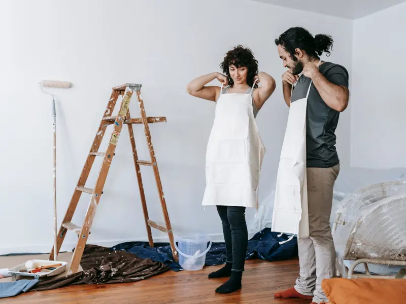 A couple prepares for home renovation, wearing aprons in an indoor setting.