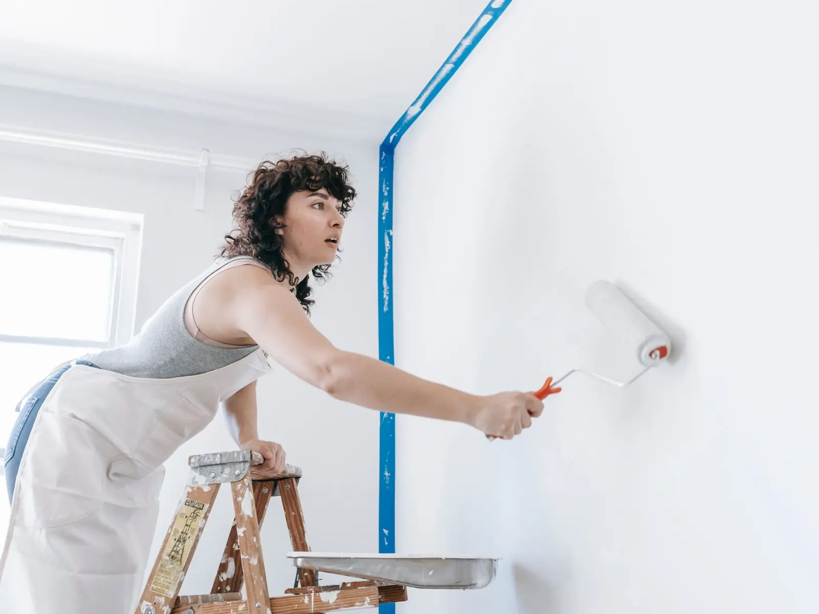 A woman stands on a ladder painting a wall white, part of a DIY home renovation.