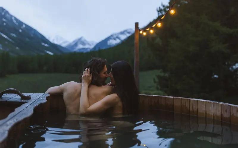 A couple embracing in a wooden hot tub with a mountain view at twilight.