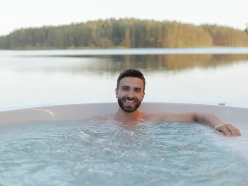 Shirtless man smiling in an outdoor hot tub by a serene lake, enjoying relaxation and wellness.