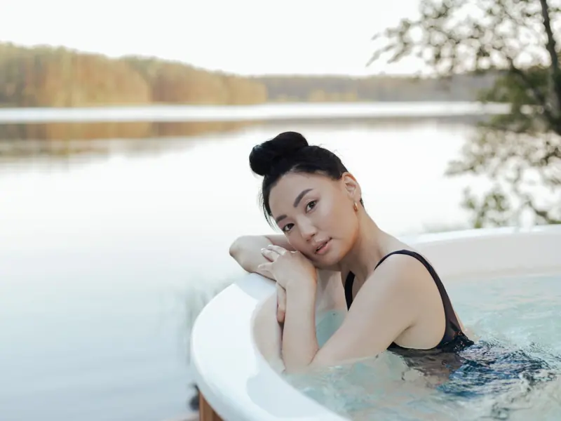 A woman enjoys a relaxing time in an outdoor hot tub by a serene lakeside setting.