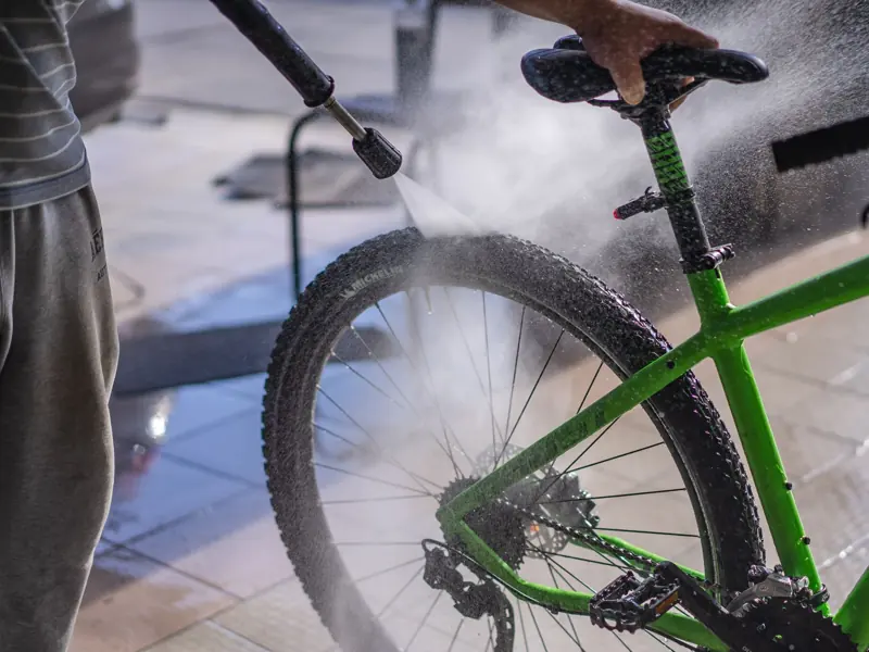 A man cleans a green bicycle with a hose, focusing on the wheel and frame outdoors.