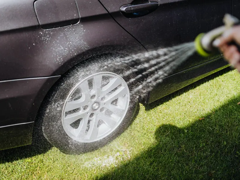Close-up shot of a car being washed, focusing on the wheel rim with water spray outdoors on grass.