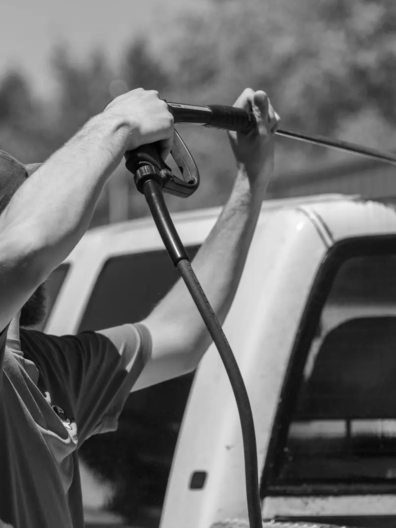 Black and white image of a man using a power spray on a truck outdoors.