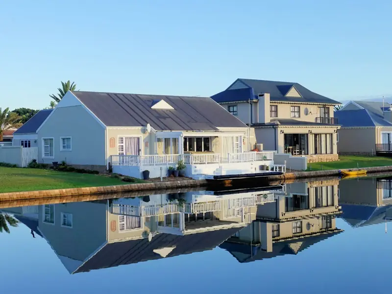 Luxury modern homes reflecting on a peaceful, clear lake during daylight, showcasing architectural beauty.