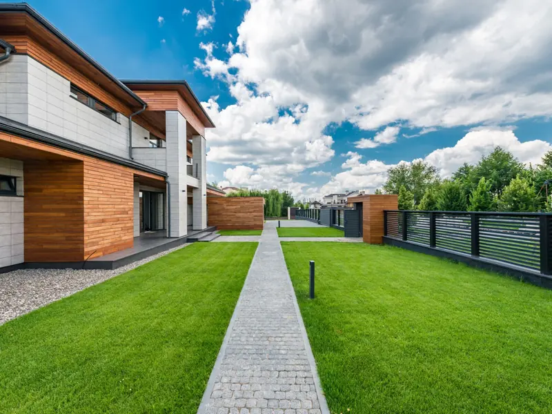 Scenery green yard of modern residential house with trees outside fence under blue cloudy sky